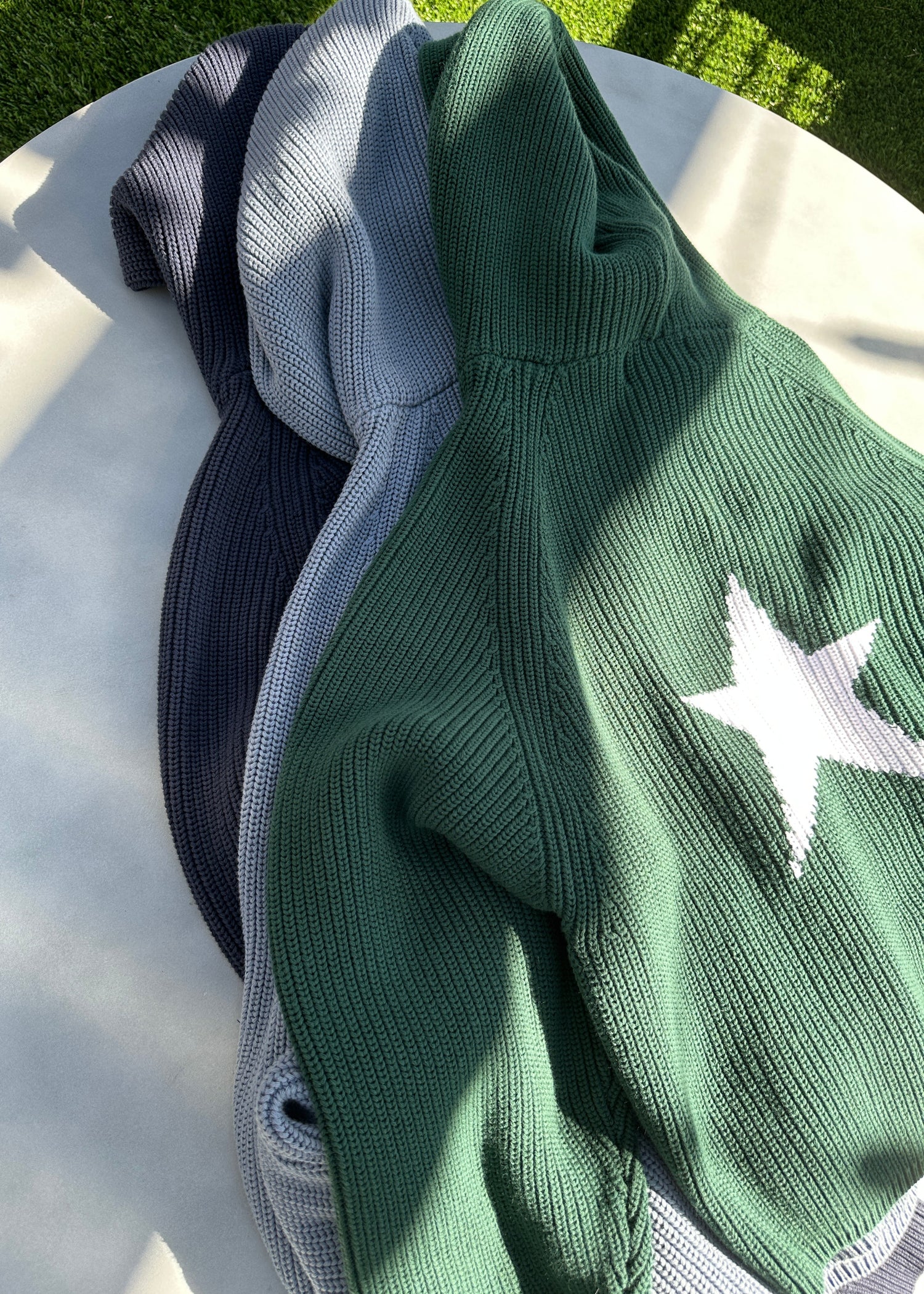 Stack of green, gray, and navy knit hoodies on a white surface with grass in the background